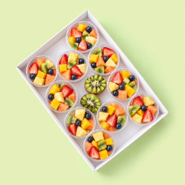 Box of 10 fresh fruit salad pots with strawberries, blueberries, kiwi fruit, mango, pineapple and melon, with two carved kiwi halves, in a white catering box on a pale green background.