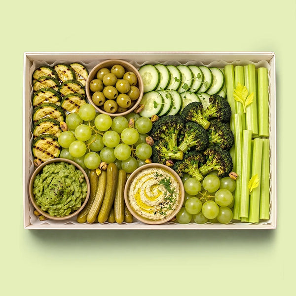Green Goddess Healthy Board in a white box with grilled zucchini, green olives, cucumber slices, broccoli florets, celery sticks, green grapes, cornichons, pistachios, guacamole and hummus.