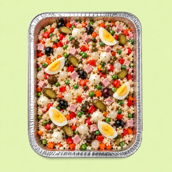 A top-down view of a prepared stuffed rice salad in a rectangular aluminum foil tray, set against a solid pale green background. The salad contains rice, green peas, red bell peppers, black olives, pickles, white feta cheese, sliced hard-boiled eggs, and pieces of ham.