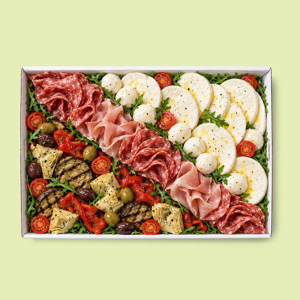 A top-down view of a rectangular charcuterie board gift box filled with sliced salami, prosciutto, mozzarella, tomatoes, grilled crackers, olives, and herbs on a pale green background.