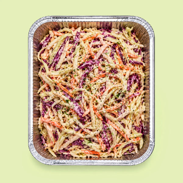 A top-down view of coleslaw in a square aluminum foil tray, set against a solid pale green background.