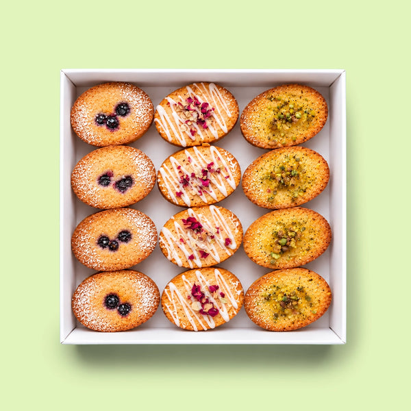 A top-down view of a white square box containing twelve assorted Friand, including meringue-dusted pastries, blueberry tarts, and pistachio-topped pastries, set against a solid pale green background.