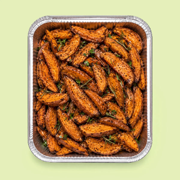 Roasted sweet potato wedges seasoned with herbs and spices, presented in a square aluminum foil pan on a light green background.