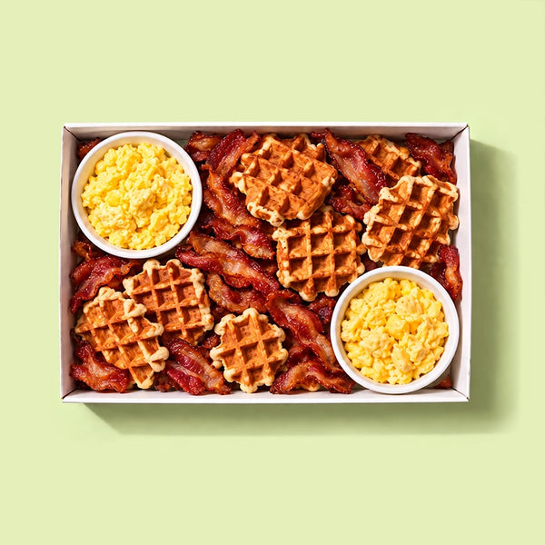 Waffle, bacon and egg platter in a white catering box with golden mini waffles, crispy bacon rashers and two pots of scrambled eggs, on a pale green background.