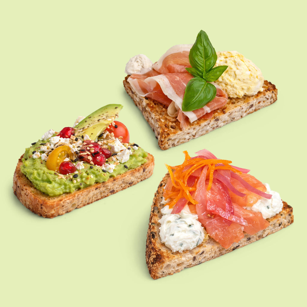 Slices of sourdough bread topped with avocado, smoked salmon, prosciutto, egg and cheese, styled as an office catering platter for morning or afternoon tea.
