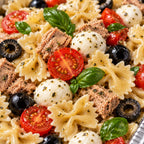 A close-up of a pasta salad in a silver foil pan, featuring bowtie pasta mixed with tuna, mozzarella balls, halved cherry tomatoes, black olives, and fresh basil leaves.