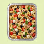 A top-down view of a prepared pasta salad in a rectangular aluminum foil tray, featuring bowtie pasta, sliced tomatoes, mozzarella balls, black olives, tuna, and fresh basil leaves on a pale green background.
