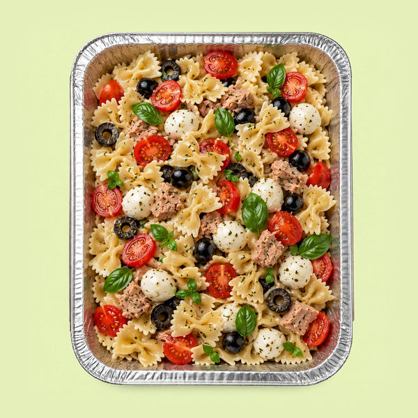 A top-down view of a prepared pasta salad in a rectangular aluminum foil tray, featuring bowtie pasta, sliced tomatoes, mozzarella balls, black olives, tuna, and fresh basil leaves on a pale green background.
