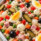 A close-up overhead view of a prepared salad or rice dish in a metal container, featuring white rice mixed with sliced hard-boiled eggs, diced pink ham, black olives, green peas, red bell peppers, orange carrots, and pickled green cucumbers.