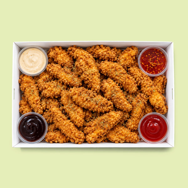 A top-down view of golden-brown crispy fried chicken tenders in a white cardboard box, served with four small plastic cups of dipping sauces on a pale yellow-green background.