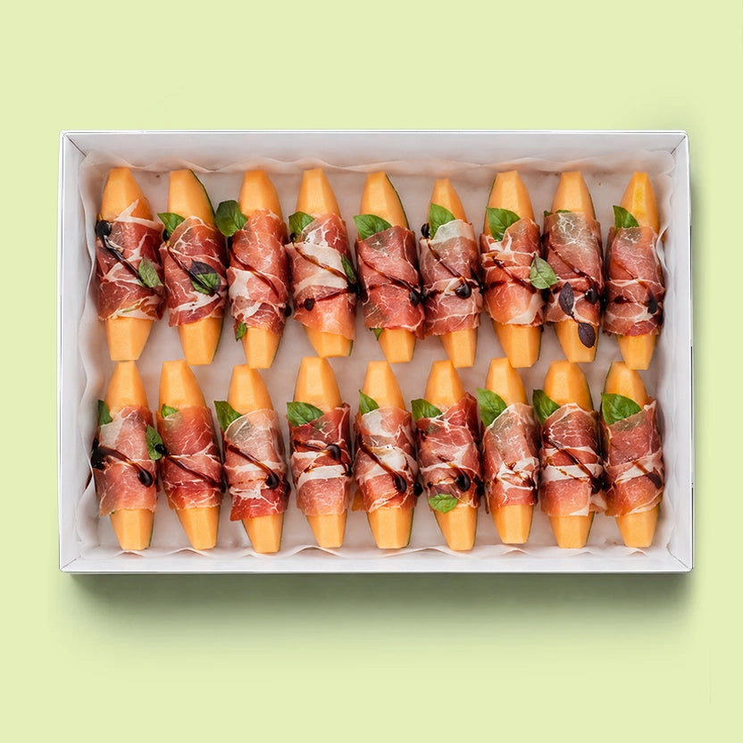 Rockmelon and prosciutto platter in a white box with each piece of rockmelon wrapped in prosciutto, topped with fresh basil leaves and a drizzle of balsamic glaze.