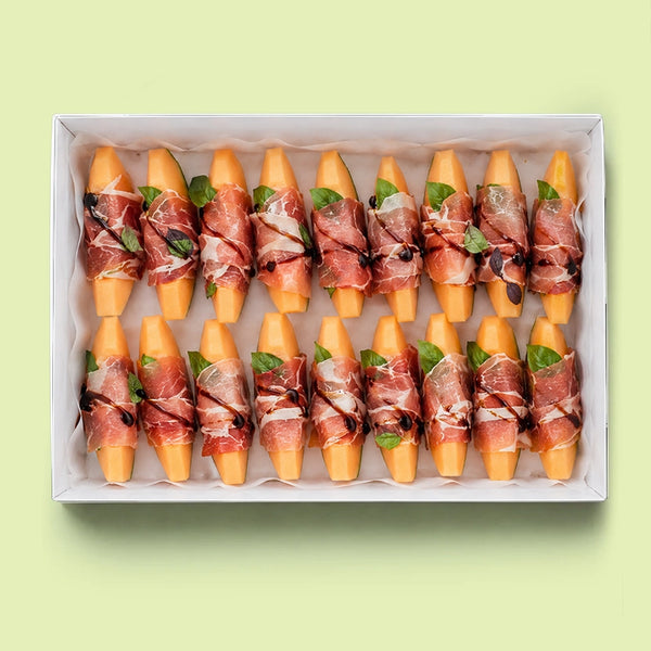 Rockmelon and prosciutto platter in a white box with each piece of rockmelon wrapped in prosciutto, topped with fresh basil leaves and a drizzle of balsamic glaze.