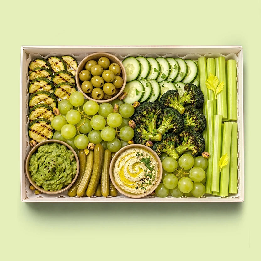 Green Goddess Healthy Board in a white box with grilled zucchini, green olives, cucumber slices, broccoli florets, celery sticks, green grapes, cornichons, pistachios, guacamole and hummus.