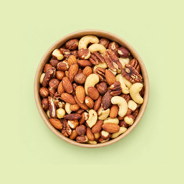 A top-down view of a mixed nut blend in a round brown bowl, set against a solid pale green background.