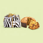 A packaged loaf of Italian panettone next to a sliced piece showing its golden-brown crumb with dark chocolate chips and tiramisu cream on a pale green background. The loaf is wrapped in black and white zebra print paper with a gold ribbon and a circular logo reading 'Muzzi'.