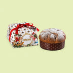 A Muzzi Italiani Panettone cake next to its decorated gift box on a pale yellow-green background. The cake is topped with powdered sugar and sits in a brown patterned paper liner, while the box features a festive holiday design with a red ribbon.