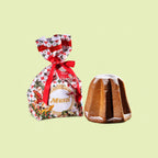 A festive packaged food product on a pale yellow-green background, showing a wrapped Muzzi candy cane-shaped treat tied with a red ribbon next to an unwrapped brown candy cane with white powdered sugar dusting.