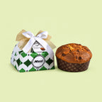 A Muzzi Pear and Chocolate Panettone cake shown next to its decorated gift box on a solid pale yellow-green background. The box is white and green with a diamond pattern and tied with a white ribbon and gold bow.
