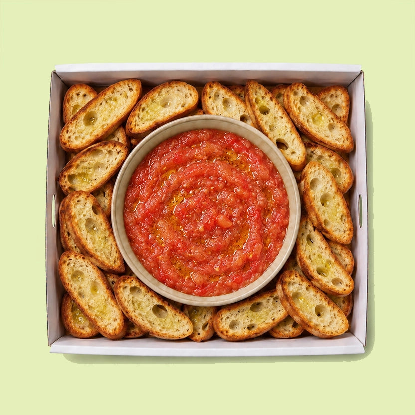 Bowl of freshly grated tomato with olive oil, surrounded by golden toasted olive oil crostini in a catering box.