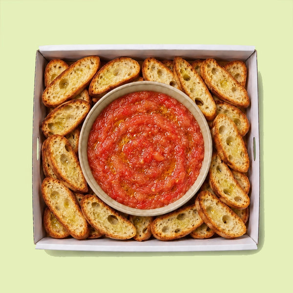 Bowl of freshly grated tomato with olive oil, surrounded by golden toasted olive oil crostini in a catering box.