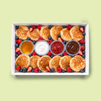 A top-down view of a white cardboard box filled with mini pancakes, fresh blueberries, strawberries, raspberries, and small containers of syrup, cream, and jam on a pale green background.