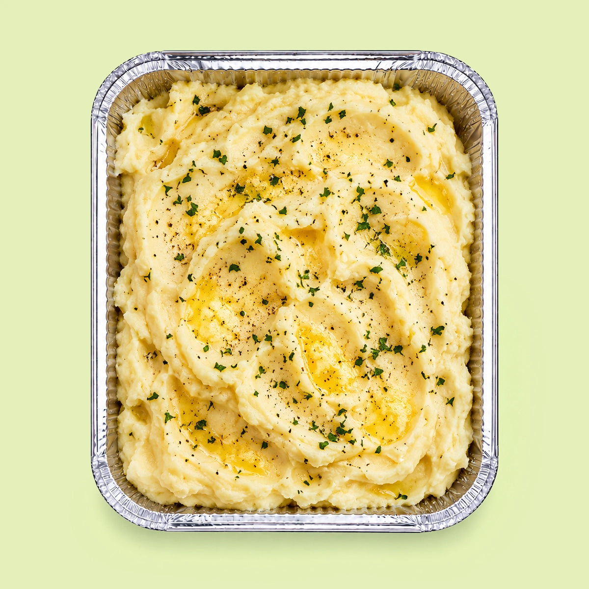 A top-down view of creamy mashed potatoes in a square aluminum foil container, garnished with chopped green herbs and a drizzle of oil, set against a solid pale green background.