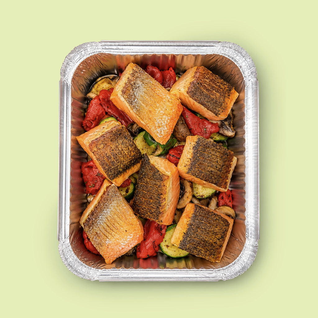 Top-down view of EATALO’s crispy-skin roast salmon platter with roasted vegetables in a catering tray on a light pink background.
