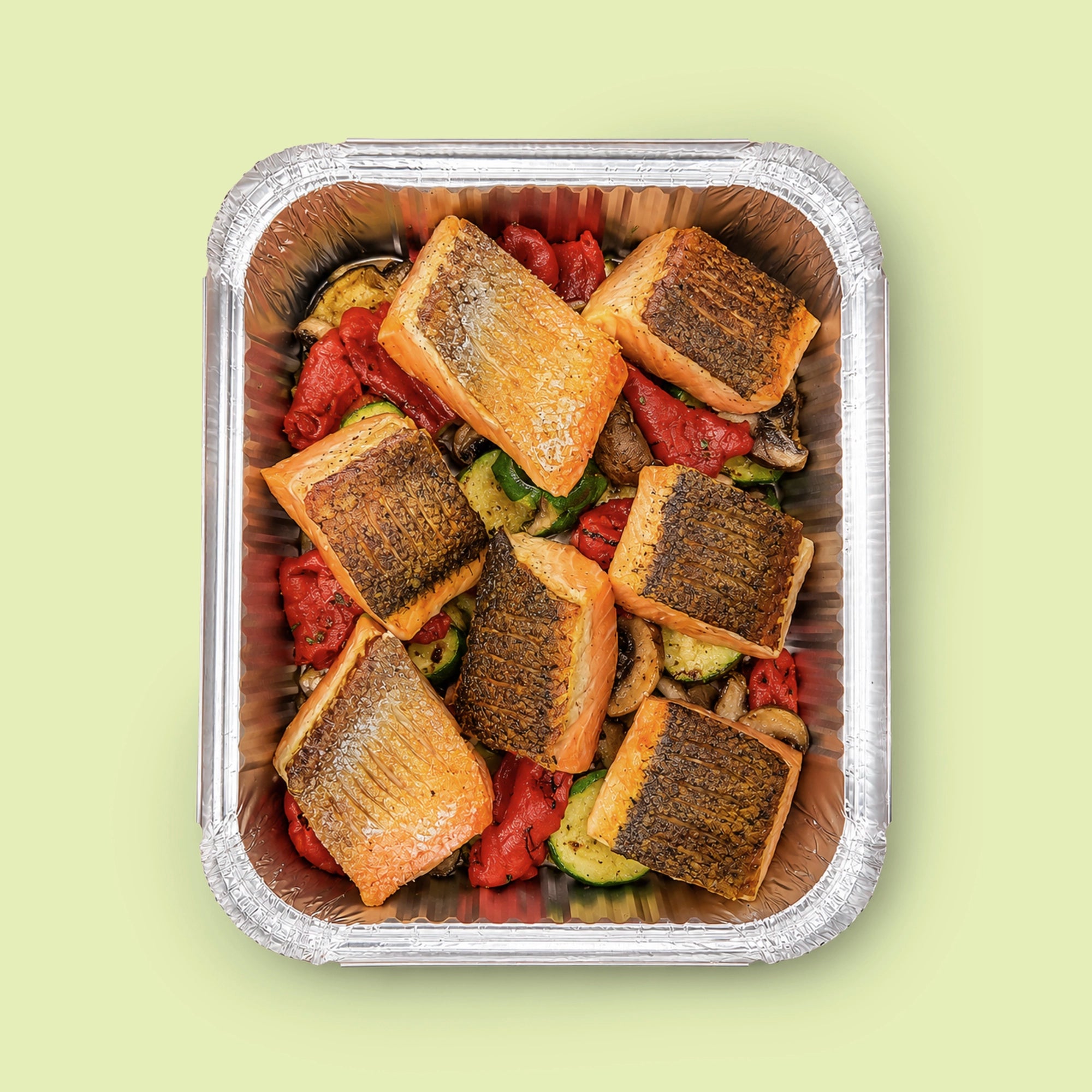 Top-down view of EATALO’s crispy-skin roast salmon platter with roasted vegetables in a catering tray on a light pink background.
