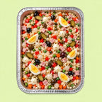 A top-down view of a prepared stuffed rice salad in a rectangular aluminum foil tray, set against a solid pale green background. The salad contains rice, green peas, red bell peppers, black olives, pickles, white feta cheese, sliced hard-boiled eggs, and pieces of ham.