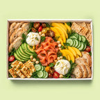 A top-down view of a gourmet charcuterie board in a white rectangular box, featuring sliced baguette, crackers, sliced cucumbers, yellow fruit, smoked salmon, mozzarella cheese, and tomatoes on a light green background.