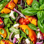 A close-up of a fresh salad with grilled peach slices, mixed greens, cherry tomatoes, red onion, and white cheese cubes on a bed of green and  basil leaves.