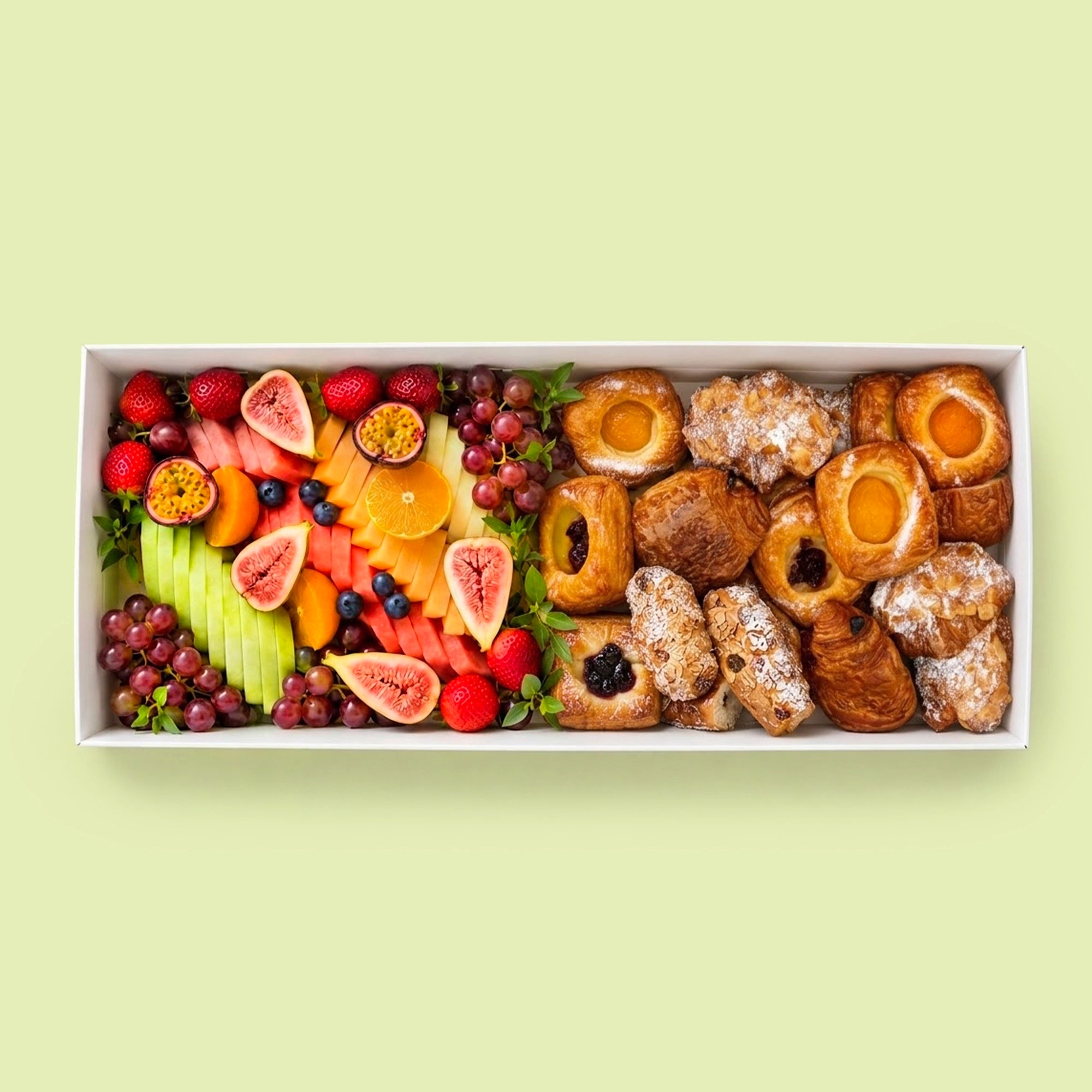 A rectangular white box filled with assorted pastries and fruit, photographed from above on a pale yellow-green background. The left side shows sliced watermelon, cantaloupe, orange, blueberries, grapes, and figs; the right side shows several golden-brown pastries, including croissants, filled pastries, and powdered-sugar-dusted treats.