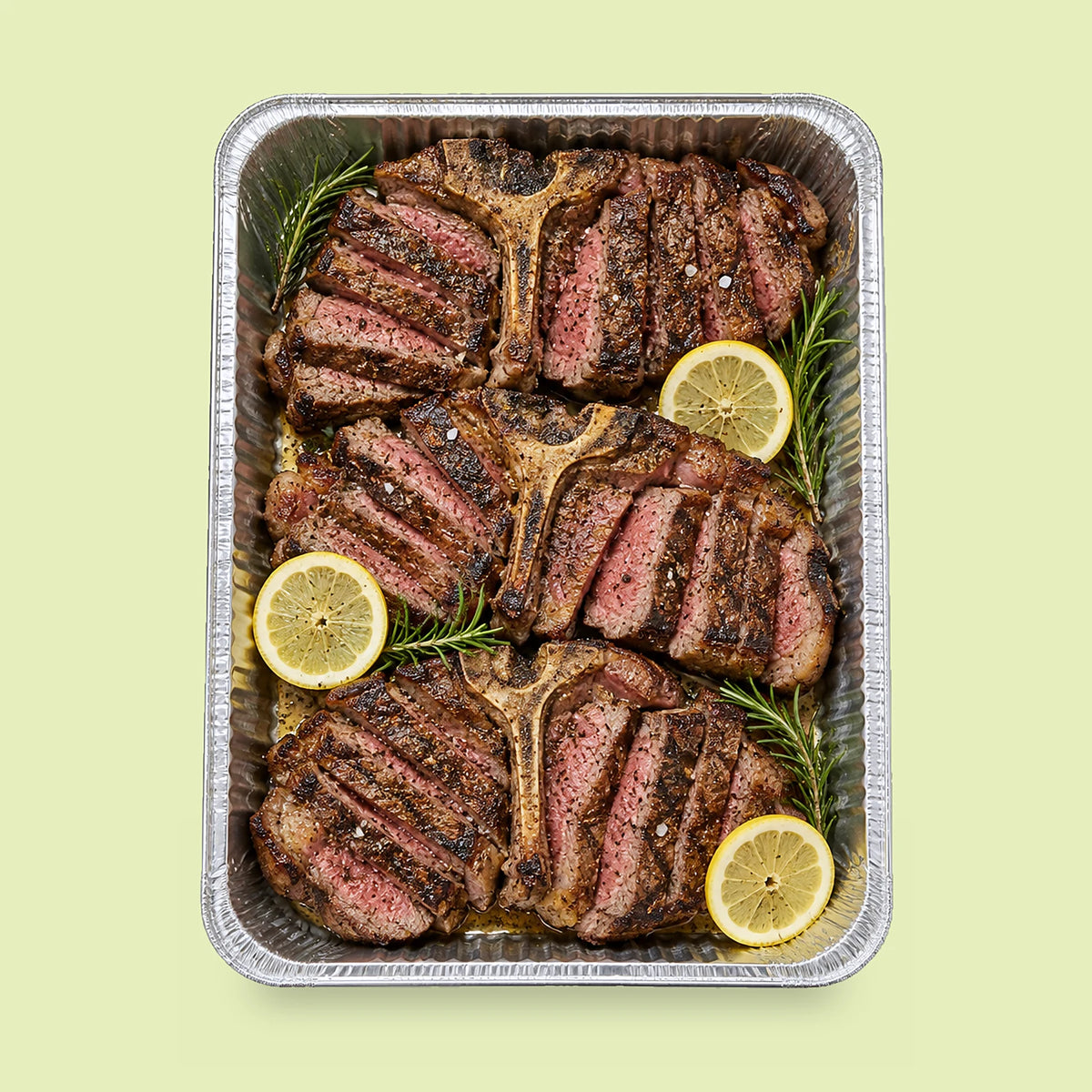 Foil catering tray of three grilled T-bone steaks, sliced off the bone, with fresh rosemary sprigs and lemon slices.