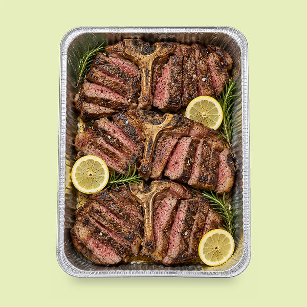 Foil catering tray of three grilled T-bone steaks, sliced off the bone, with fresh rosemary sprigs and lemon slices.