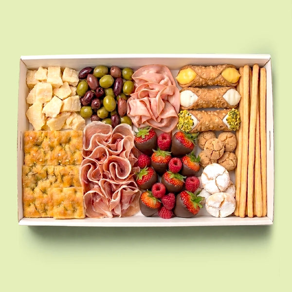 Italian mixed banquet platter in a white box with parmigiano chunks, prosciutto, mortadella, mixed olives, cannoli, chocolate-dipped strawberries, raspberries, amaretti, focaccia and grissini.
