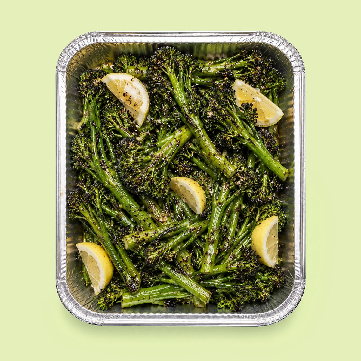 Roasted broccolini with lemon wedges in a square aluminum foil pan, presented on a light green background.