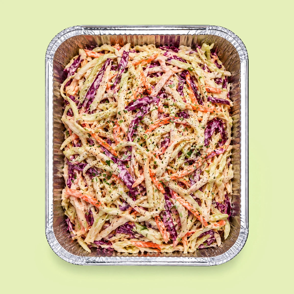 A top-down view of coleslaw in a square aluminum foil tray, set against a solid pale green background.