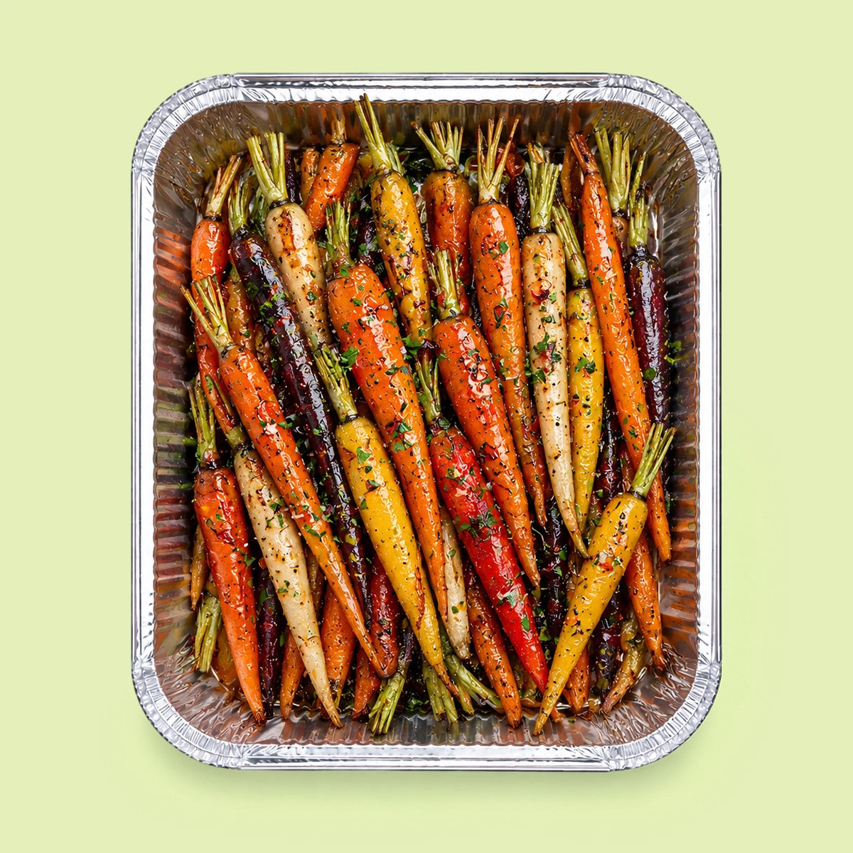 A top-down view of roasted multicolor baby carrots seasoned with herbs and spices, arranged inside a square aluminum foil tray against a pale green background.