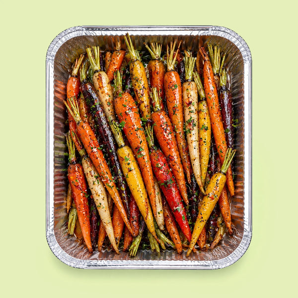 A top-down view of roasted multicolor baby carrots seasoned with herbs and spices, arranged inside a square aluminum foil tray against a pale green background.
