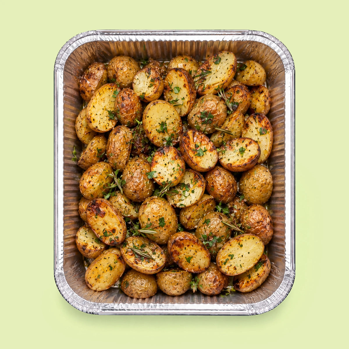 Roasted baby potatoes in a square aluminum foil tray, garnished with chopped herbs, on a solid pale green background.