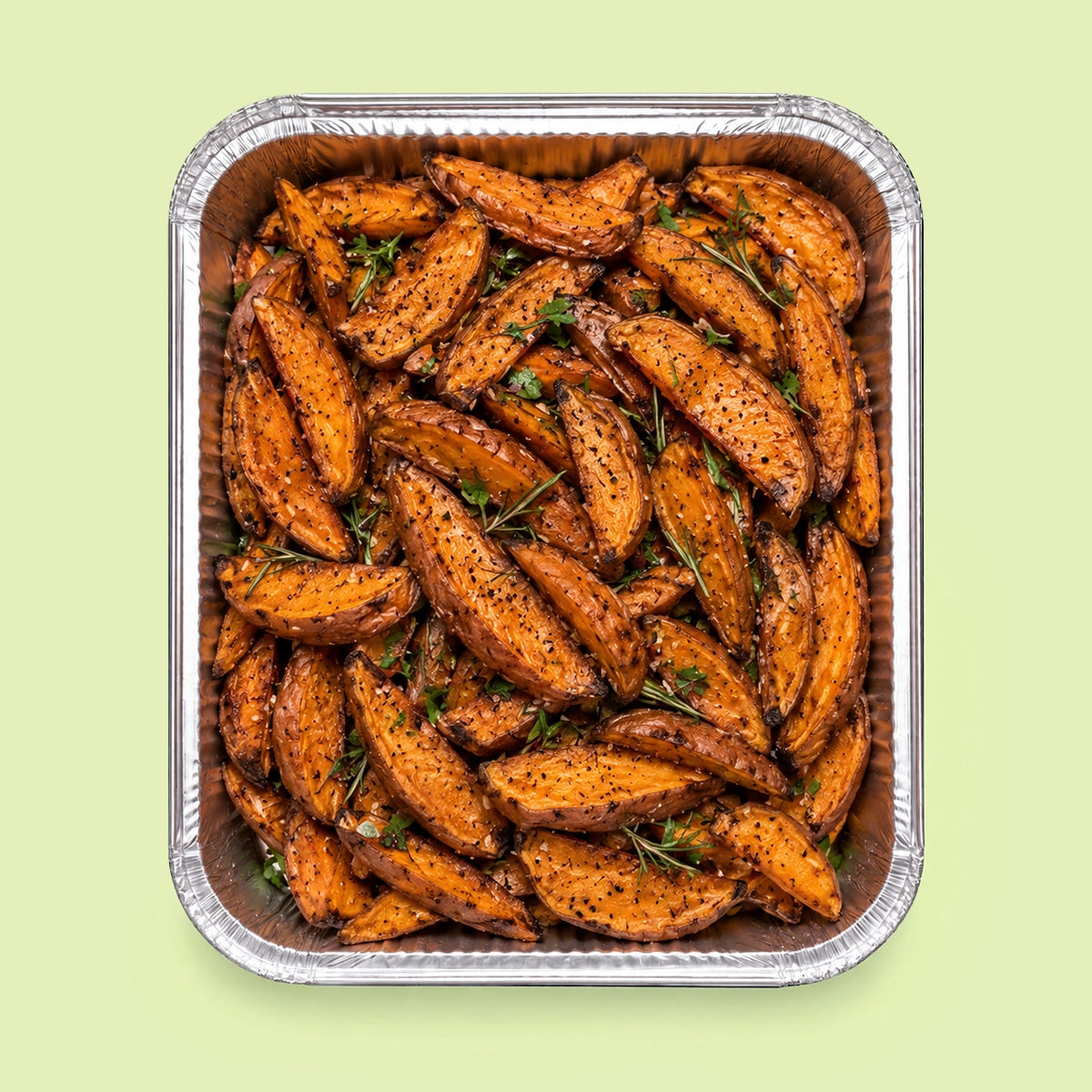 Roasted sweet potato wedges seasoned with herbs and spices, presented in a square aluminum foil pan on a light green background.