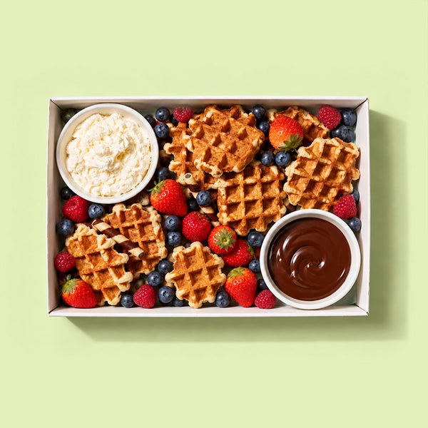 Waffle platter in a white catering box with mini waffles, strawberries, raspberries, blueberries, chocolate wafer rolls, a pot of whipped cream and a pot of chocolate, on a pale green background.