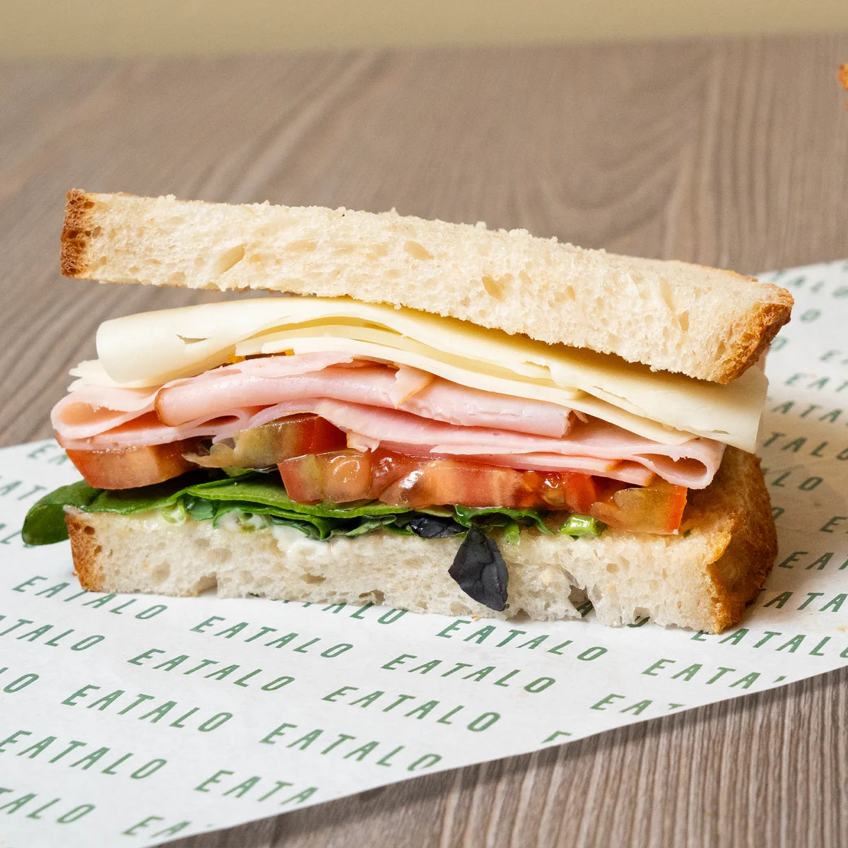 Sandwich with ham, cheese, lettuce, and tomato on a wooden surface with Eatalo branded paper.
