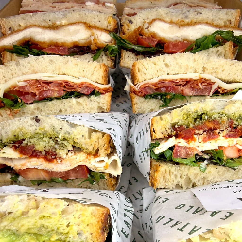 Multiple Italian sandwiches cut in half, stacked and wrapped in paper with visible text. Eatalo, in Sydney.