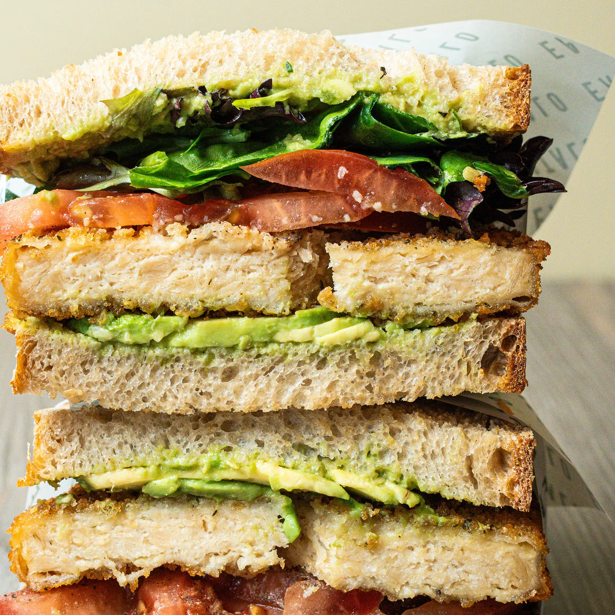 Stack of a sandwich with avocado, chicken schnitzel, tomato, and leafy greens on a light background