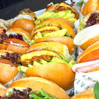 Row of small burgers with buns, lettuce, and other ingredients on a tray.