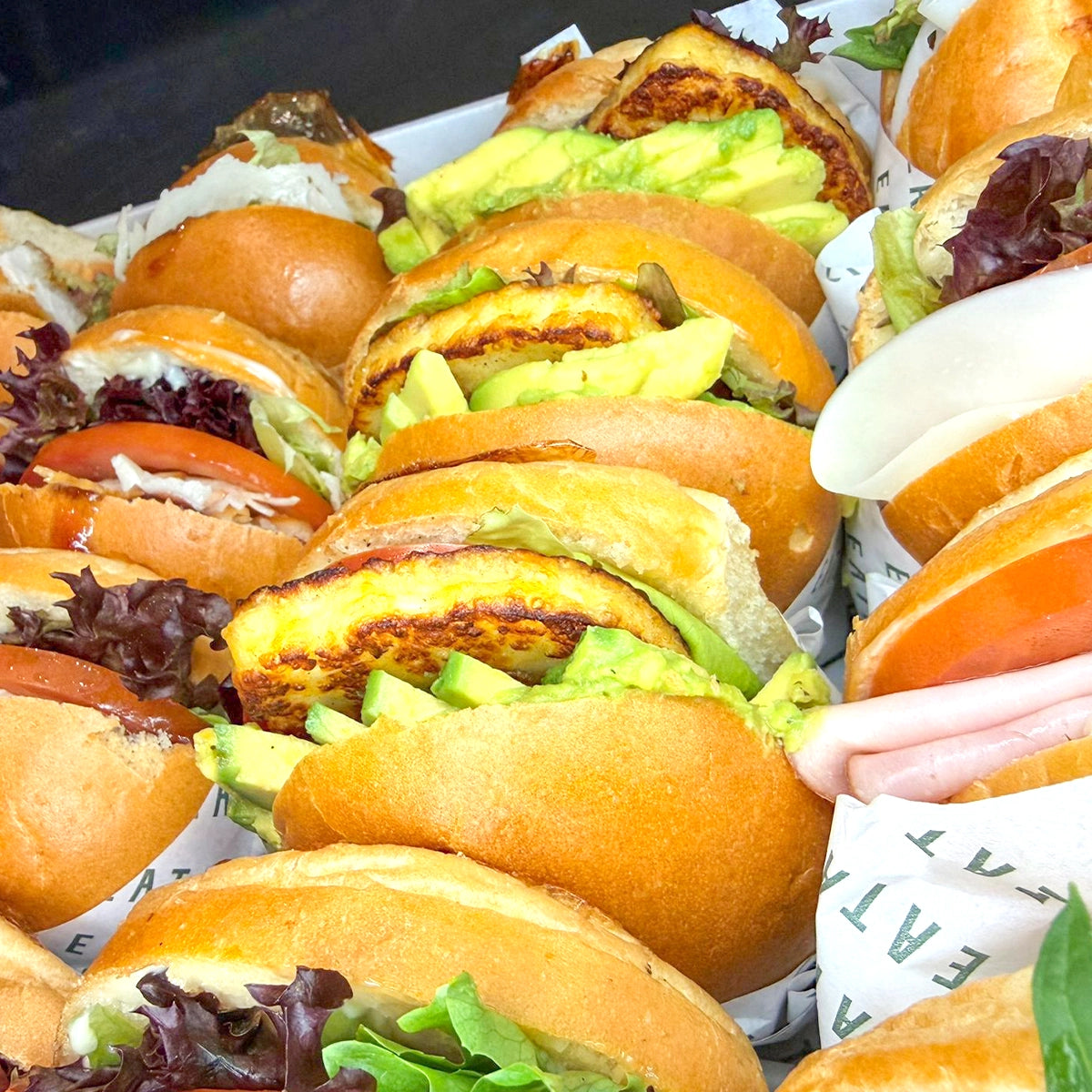 Row of small burgers with buns, lettuce, and other ingredients on a tray.