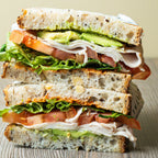 Stacked sandwich with chicken breast, lettuce, tomato, and avocado on a wooden surface.