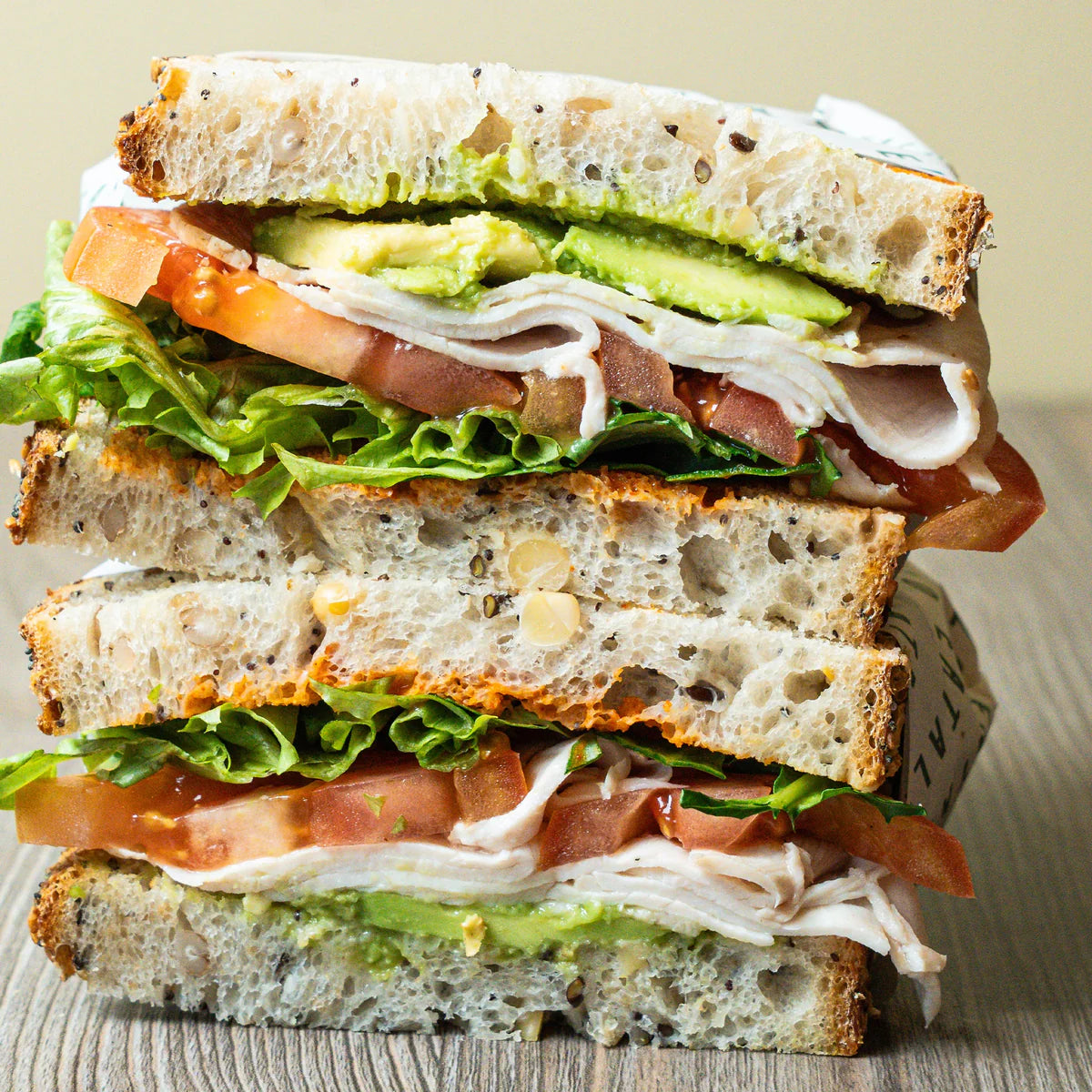 Stacked sandwich with chicken breast, lettuce, tomato, and avocado on a wooden surface.