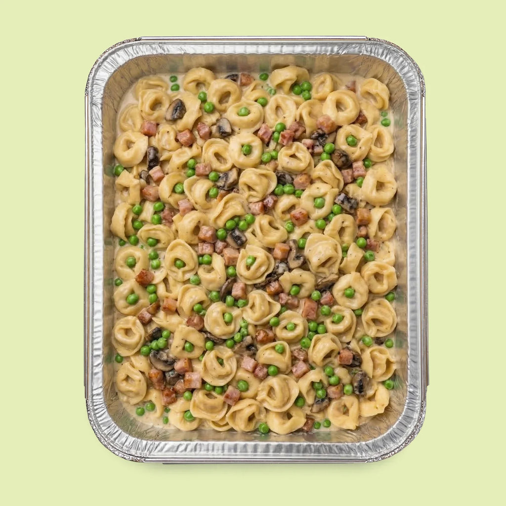 Foil pan of Italian tortellini pasta with peas and ham on a light green background